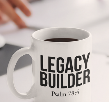 White mug with 'Legacy Builder Psalm 78:4' text on a desk with blurred hands and laptop in the background