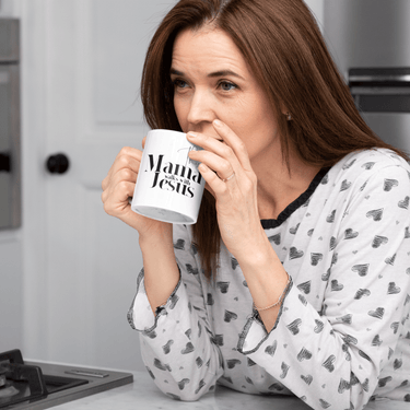 Woman in pajamas holding a mug with 'Mama de Jesus' text in a kitchen.