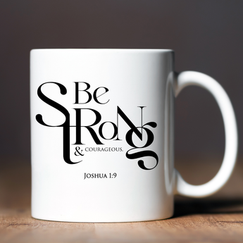 White mug with 'Be Strong & Courageous' text on a wooden surface