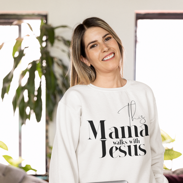Woman wearing a white sweatshirt with 'This Mama Walks with Jesus' text, holding a laptop in a home setting.